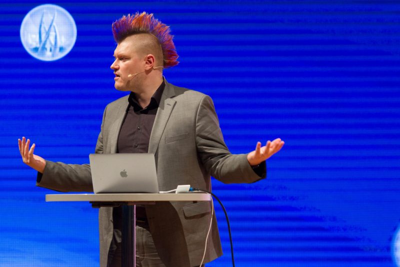 Redner mit bunter Irokesenfrisur spricht auf der Bühne, Laptop auf Pult, blaue Hintergrundbeleuchtung, Kongress und Technologieveranstaltung