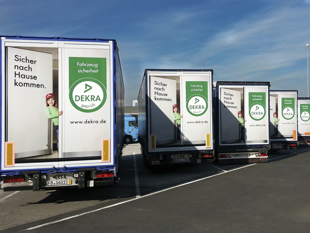 Reihe von LKW mit DEKRA-Werbeflächen und dem Slogan „Sicher nach Hause kommen“ auf einem Parkplatz
